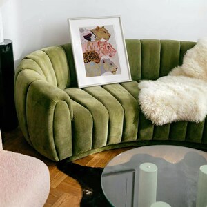 May include: A curved olive green velvet sofa with vertical channel tufting. A white framed print featuring four stacked stylised big cats in shades of tan, pink, brown, and blue rests on the seat. A fluffy white sheepskin throw lies on the right.
