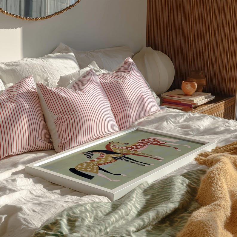May include: A framed art print featuring three stylized horses in motion, with a light green background. The horses have patterned coats in red, black, and gold. The print is displayed on a bed with white linens and red striped pillows.