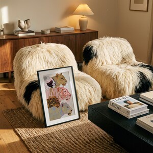 May include: A framed rectangular art print featuring four stacked stylised big cat illustrations in shades of tan, pink, brown, and blue, positioned in front of two cream-coloured, long-haired faux fur armchairs with black patches.