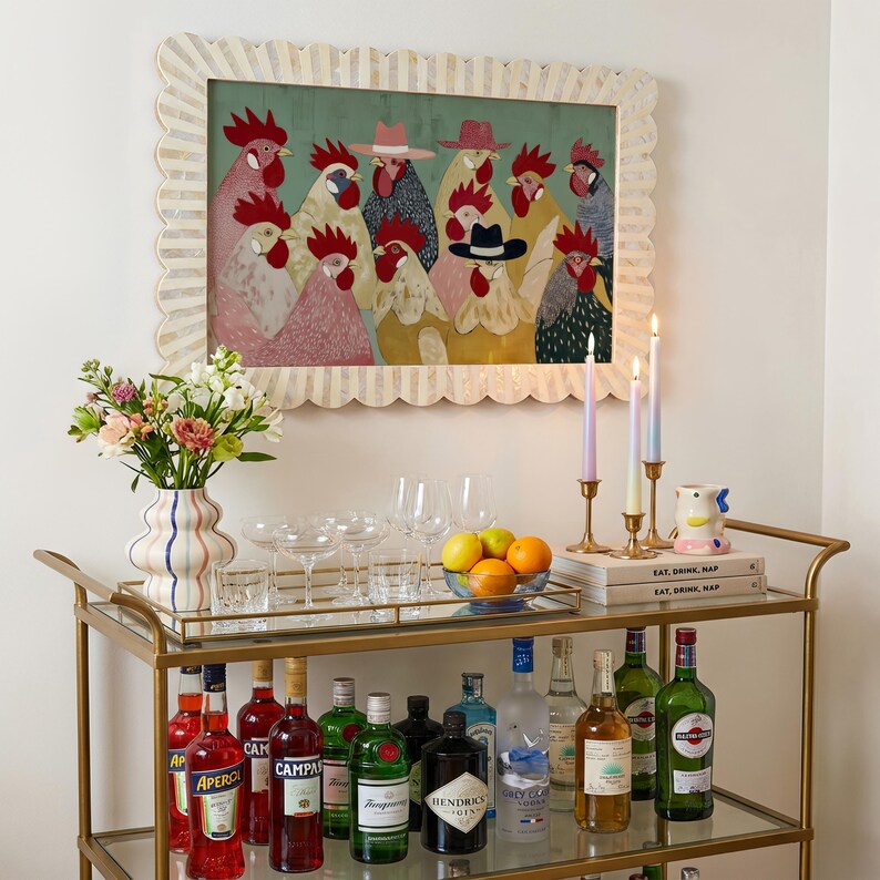 K&ouml;nnte beinhalten: Ein goldener Servierwagen mit Glasplatte, auf dem verschiedene Spirituosenflaschen, Gl&auml;ser und eine Blumenvase stehen. Dar&uuml;ber h&auml;ngt ein gerahmtes Kunstwerk mit H&uuml;hnern, die H&uuml;te tragen. Kerzen und B&uuml;cher sind ebenfalls zu sehen.