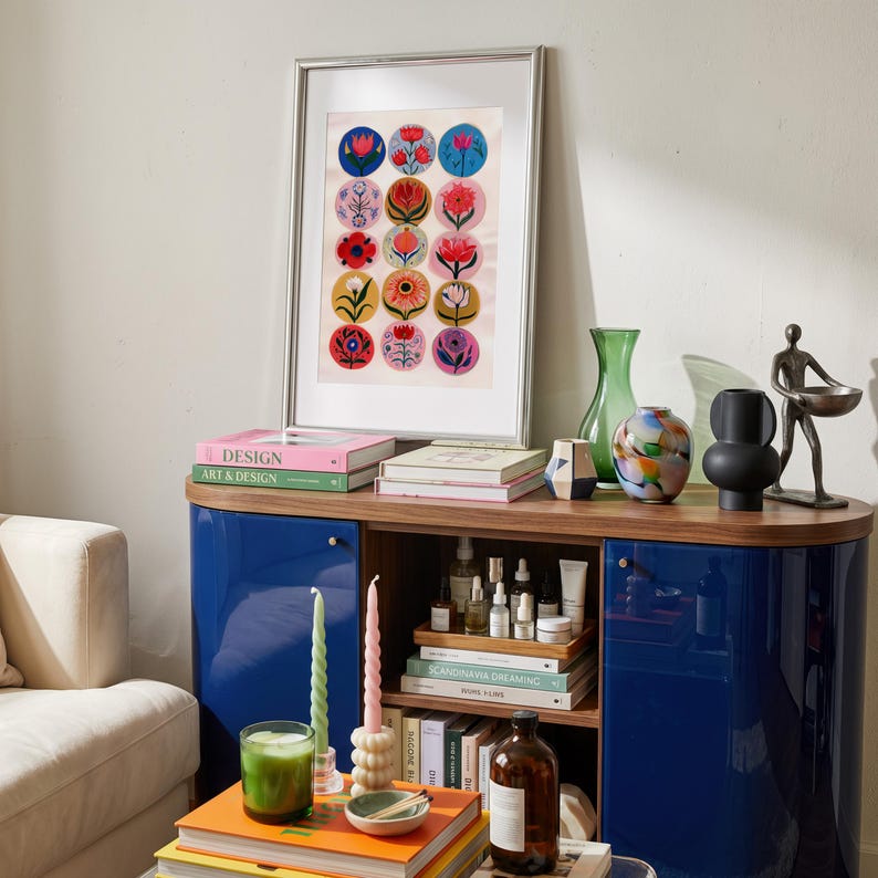 May include: A framed artwork featuring colorful floral designs, displayed on a blue cabinet with a wooden top. The cabinet holds books, candles, and decorative vases. A green candle and a stack of books are on a table in front.