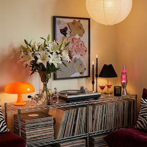 May include: A metal wire shelving unit holds a record player, a glass vase with white lilies, an orange mushroom lamp, a lava lamp, two black taper candles, a small black pleated lamp, and stacks of vinyl records. A framed lion print hangs above.