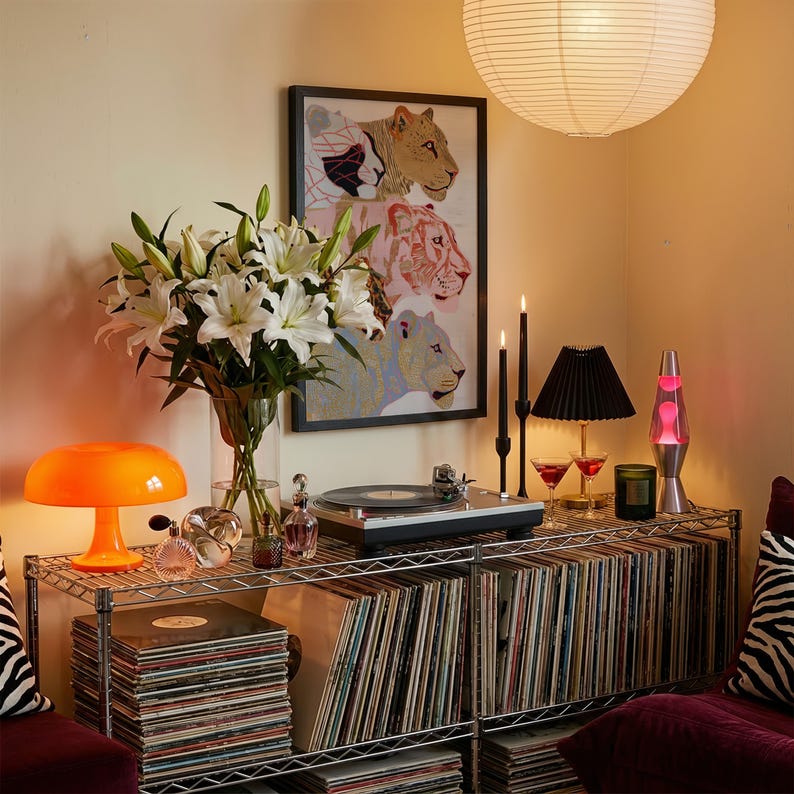 May include: A metal wire shelving unit holds a record player, a glass vase with white lilies, an orange mushroom lamp, a lava lamp, two black taper candles, a small black pleated lamp, and stacks of vinyl records. A framed lion print hangs above.