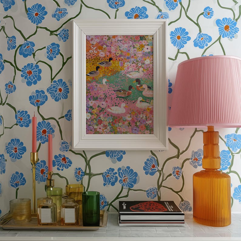 May include: A framed painting of ducks in a floral landscape hangs on a wall with a blue floral pattern. A pink lampshade sits atop an orange lamp. A tray holds candles, perfume bottles, and a green glass.