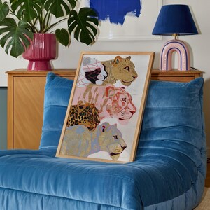 May include: A rectangular framed art print featuring a vertical stack of four stylised big cats. The illustration includes a white and black cat, a tan lion, a pink tiger, and a blue leopard, all rendered with intricate line work and patterns.