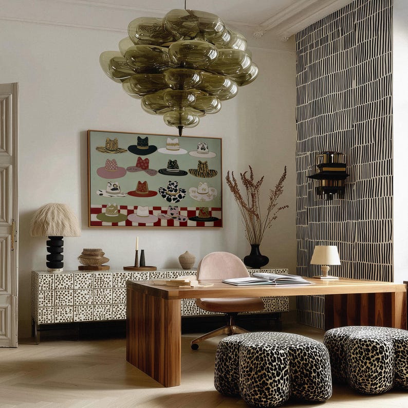 May include: An interior design scene featuring a modern office space. A large, olive-green glass chandelier hangs above a wooden desk and two leopard-print ottomans. A painting of hats hangs on the wall, and a patterned cabinet is visible.
