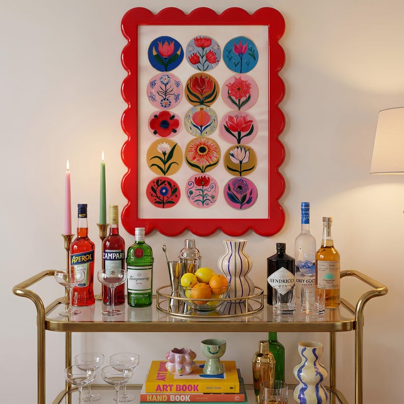 May include: A red scalloped-edge framed artwork with colorful floral designs hangs above a gold bar cart. The cart holds bottles of liquor, glassware, a cocktail shaker, and a bowl of citrus fruit. Lit candles add ambiance.