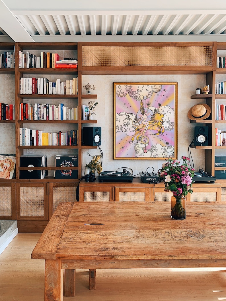 May include: A wooden table with a light wood finish sits in front of a wall with a bookshelf, a record player, and a framed print of a woman riding a lion. The print has a pink, purple, and yellow color scheme. There are flowers in a vase on the table.