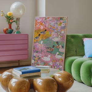 May include: A framed artwork depicting ducks in a floral setting. The painting is in a light wood frame, leaning against a green velvet sofa. A pink chest of drawers, a round wooden coffee table, and a decorative lamp are also in view.