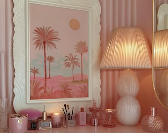 May include: A pink and white vanity scene featuring a framed landscape painting of palm trees, a pleated lampshade, and various beauty products. The painting is in a white scalloped frame. The scene includes a candle, perfume bottles, and makeup brushes.