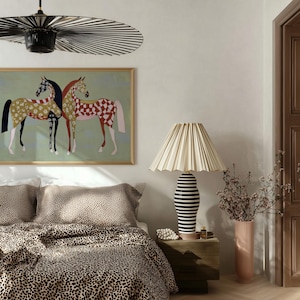 May include: A bedroom scene featuring a bed with leopard print bedding, a framed painting of two horses, and a table lamp with a pleated lampshade. The lamp has a black and white striped base. A tall pink vase holds dried flowers.
