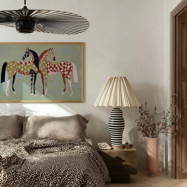 May include: A bedroom scene featuring a bed with leopard print bedding, a framed painting of two horses, and a table lamp with a pleated lampshade. The lamp has a black and white striped base. A tall pink vase holds dried flowers.