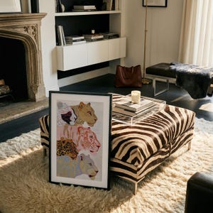 May include: A rectangular framed wall art piece featuring a vertical stack of four stylised big cat illustrations in shades of gold, pink, brown, and blue, set within a simple black frame and displayed on a white shaggy rug in a room.