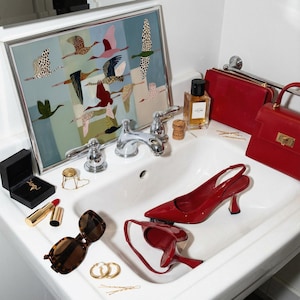 May include: A white bathroom sink containing a pair of red patent leather slingback heels, a red handbag, a red clutch, a bottle of Celine perfume, brown tortoiseshell sunglasses, gold hoop earrings, a lipstick, and a framed print of flying birds.