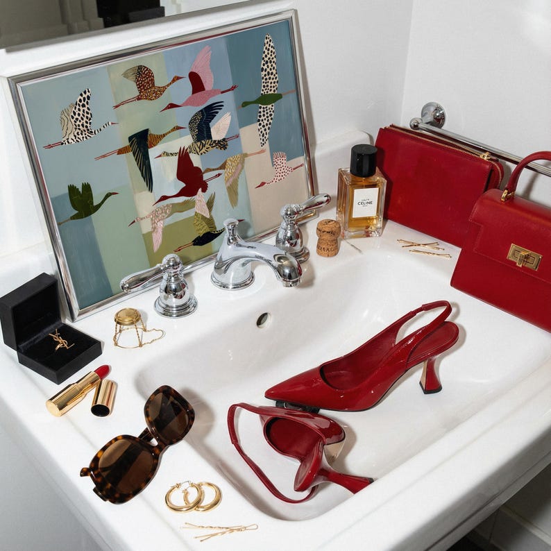 May include: A white bathroom sink containing a pair of red patent leather slingback heels, a red handbag, a red clutch, a bottle of Celine perfume, brown tortoiseshell sunglasses, gold hoop earrings, a lipstick, and a framed print of flying birds.