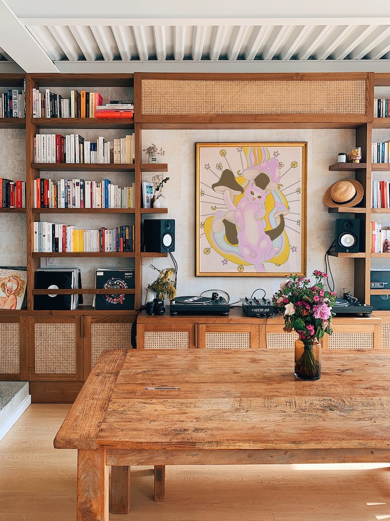 Puede incluir: Una mesa de madera con un acabado de madera clara se encuentra frente a una pared con una estanter&iacute;a, un tocadiscos y una impresi&oacute;n enmarcada de un gato rosa con setas. La impresi&oacute;n tiene un marco dorado y un fondo blanco. La estanter&iacute;a est&aacute; llena de libros y tiene un panel de rat&aacute;n tejido encima. El tocadiscos est&aacute; sobre un gabinete de madera con un panel de rat&aacute;n tejido.