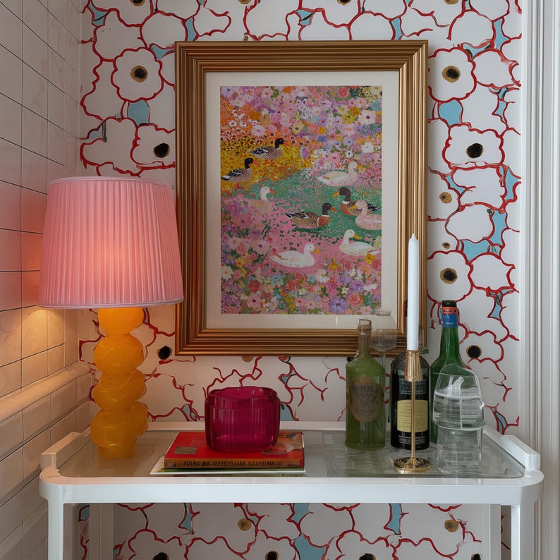 May include: A decorative display with a framed floral print, a pink lampshade on a yellow lamp, and a small table with a candle holder, bottles, and a pink glass. The wallpaper features a white floral pattern.