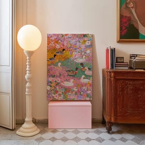 May include: A decorative white floor lamp with a globe-shaped light stands beside a painting of ducks in a floral setting. A pink box is beneath the painting, and a wooden cabinet is to the right. The painting is a vibrant display of colour.