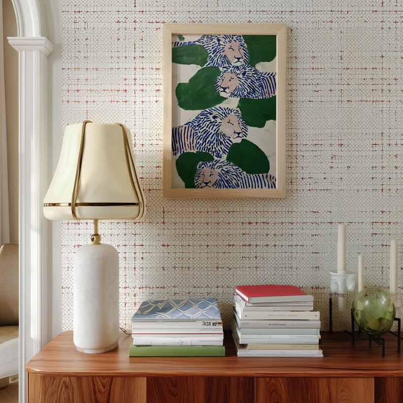 May include: A decorative scene featuring a wooden cabinet with a marble lamp and a framed artwork of lions. The artwork is above a stack of books. The lamp has a cream-colored shade with gold trim. The background is a patterned wallpaper.