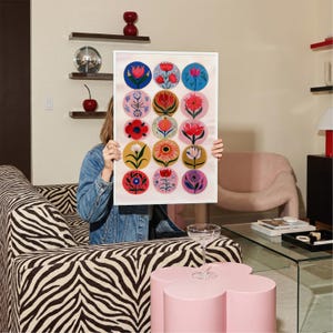 May include: A framed art print featuring a collection of colorful floral designs in circular patterns. The artwork is held in front of a person, showcasing a variety of flower illustrations in shades of red, pink, blue, yellow, and white. The print is displayed in a bright, modern living room.