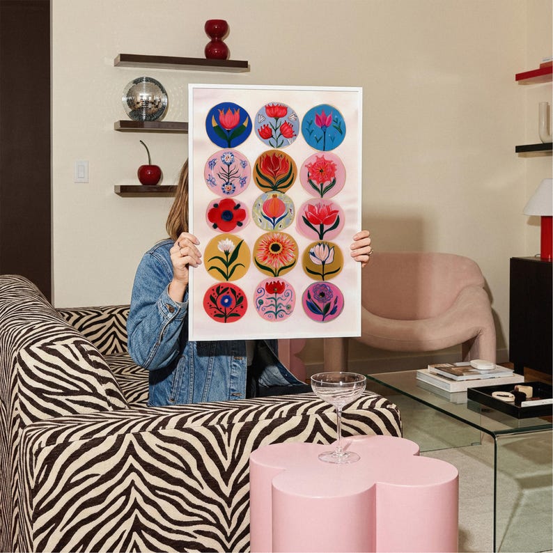May include: A framed art print featuring a collection of colorful floral designs in circular patterns. The artwork is held in front of a person, showcasing a variety of flower illustrations in shades of red, pink, blue, yellow, and white. The print is displayed in a bright, modern living room.