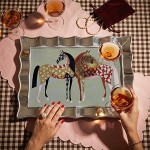 May include: A decorative mirrored tray with a painting of two horses, one with a floral pattern and the other with a checkered pattern. The tray is surrounded by glasses of rose wine, a pearl necklace, and a velvet bag.