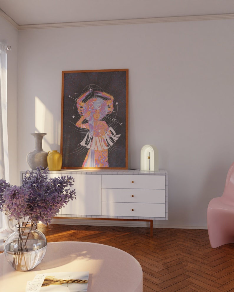 Op de afbeelding: Een witte commode met drie lades en gouden knoppen. Een paarse bloemschikking staat in een heldere glazen vaas aan de linkerkant van de commode. Een ingelijste poster met een psychedelisch ontwerp hangt aan de muur boven de commode. Een witte booglamp staat aan de rechterkant van de commode. Een roze stoel staat op de achtergrond.