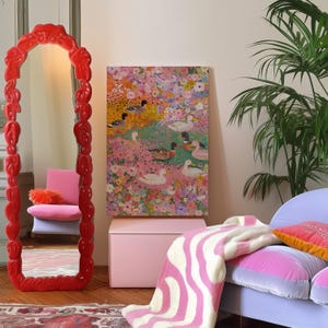 May include: A brightly coloured interior with a large red mirror with an elaborate frame. A painting of ducks in a floral setting is on the wall. A pink chair, a pink storage box, and a patterned blanket contribute to the room's overall design.