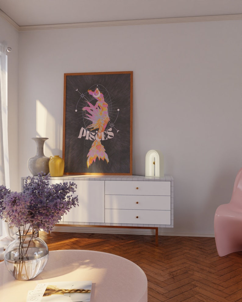 Pu&ograve; includere: Una stampa artistica incorniciata con un colorato design dei Pesci e la parola "Pisces". L'opera d'arte &egrave; appesa sopra un mobile bianco con ferramenta dorata. Una sedia rosa e un tavolino rotondo con un vaso di fiori sono in primo piano.