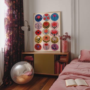 May include: A framed floral art print featuring colorful, circular designs of various flowers. The print is displayed on a wooden cabinet with books and a pink vase. A disco ball and a bed with pink bedding are also visible.
