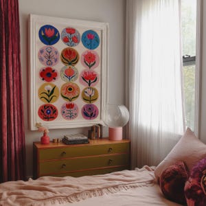 May include: A framed artwork featuring colorful floral designs in circular patterns. The artwork hangs above a green and wood-toned dresser. A pink lamp and vase add pops of color to the room, with a bed covered in a pink blanket.