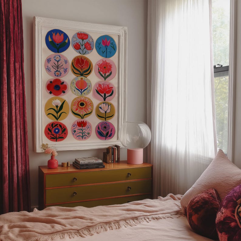 May include: A framed artwork featuring colorful floral designs in circular patterns. The artwork hangs above a green and wood-toned dresser. A pink lamp and vase add pops of color to the room, with a bed covered in a pink blanket.