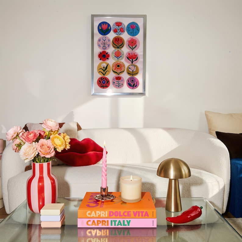 May include: A living room scene featuring a framed floral print, a white sofa, and a glass coffee table. The table holds a red and white striped vase with flowers, books, a candle, a brass lamp, and a red chili pepper.