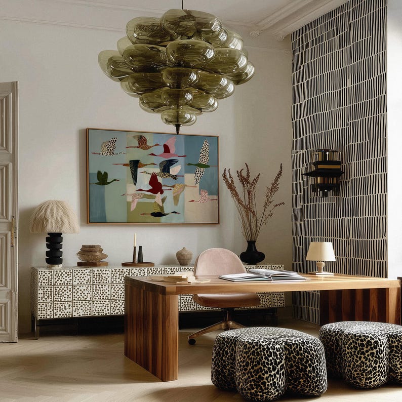 May include: A room interior featuring a large olive-green glass bubble chandelier, a rectangular painting of flying birds, a white sideboard with black dots, a wooden desk, a pink chair, and two leopard-print upholstered stools on a floor.