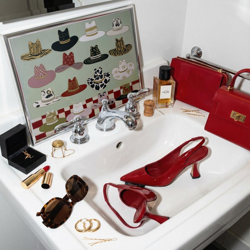 May include: A collection of fashion accessories and decor arranged in a bathroom sink. Items include a framed hat print, red handbags, a perfume bottle, red heels, sunglasses, jewelry, and lipstick.