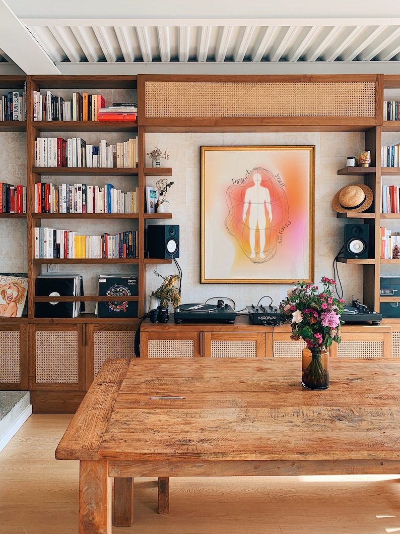 May include: A wooden table with a light wood finish sits in front of a wall with a bookshelf and a framed print that reads "Protect your energy". The print is surrounded by a light pink, orange, and yellow gradient. There is a vase of pink and purple flowers on the table.