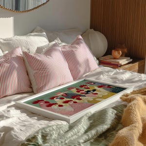 K&ouml;nnte beinhalten: Ein gerahmtes Kunstwerk mit einem bunten Hahn-Design liegt auf einem Bett mit wei&szlig;er Bettw&auml;sche und rot-wei&szlig; gestreiften Kissen. Das Kunstwerk befindet sich in einem wei&szlig;en Rahmen. Eine gelbe Decke und eine gr&uuml;n-wei&szlig; gemusterte Decke befinden sich ebenfalls auf dem Bett.