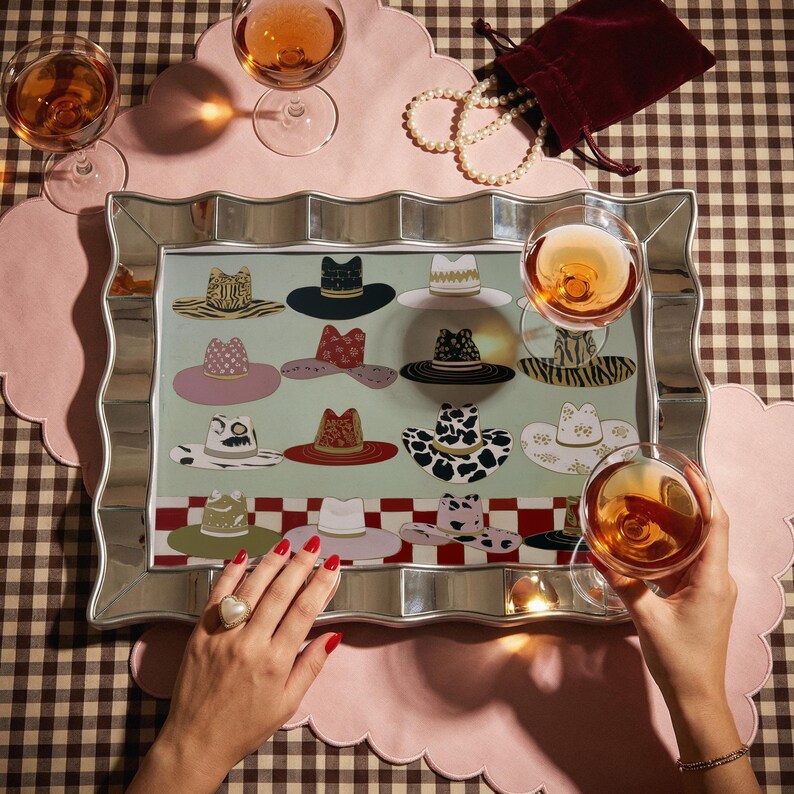 May include: A decorative mirrored tray with a collection of illustrated hats in various styles and patterns. The tray is surrounded by glasses of rose wine, a pink scalloped placemat, and a pearl necklace.