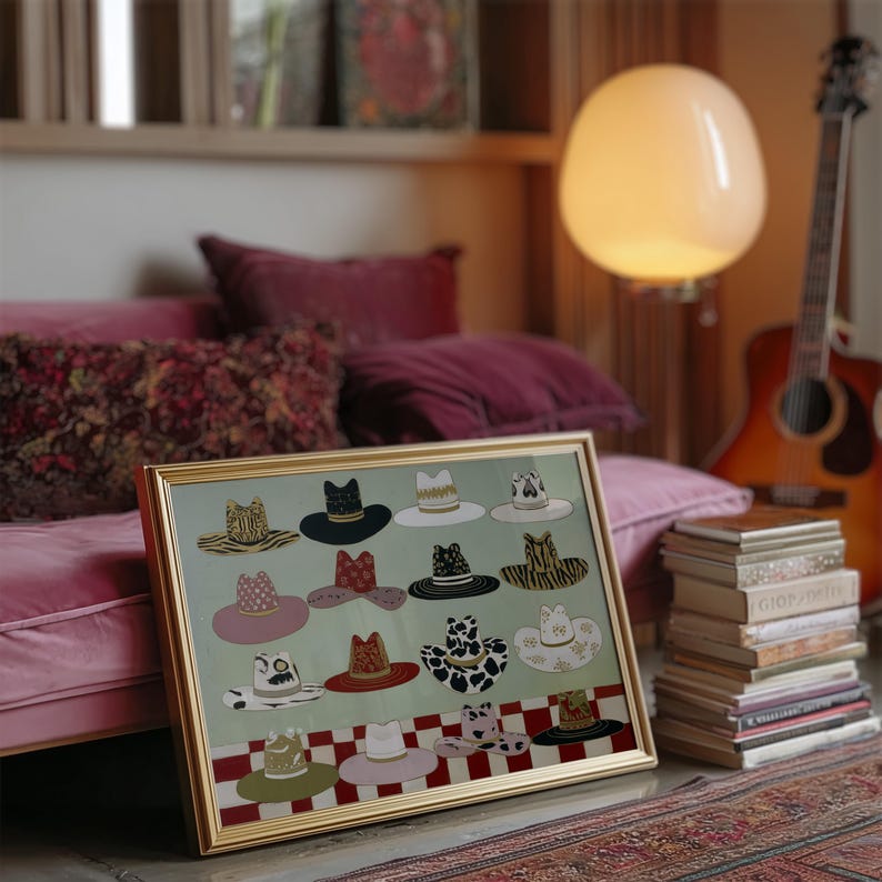 May include: A framed artwork featuring a collection of illustrated hats in various styles and patterns. The hats are displayed against a light green background with a red and white checkered border. The artwork is in a gold frame, leaning against a pink couch.