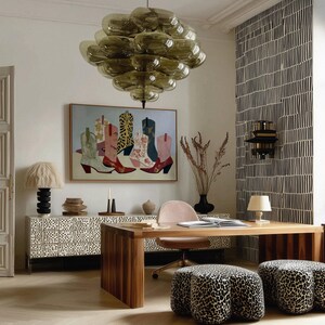 May include: An interior design scene featuring a large, abstract painting of cowboy boots in various colors. A large, olive-green glass chandelier hangs above a wooden desk and leopard-print ottomans. A patterned cabinet and black and white wallpaper complete the room.