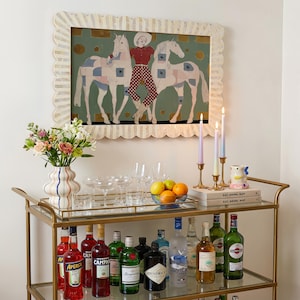 Modern western art print featuring a stylish cowgirl in a white hat and red polka dot outfit posed between two white patchwork horses. Soft sage green background with gold geometric accents and quilt-like horse patterns create a chic, editorial equestrian vibe. Perfect for contemporary western decor, ranch house interiors, cowgirl chic bedrooms, offices, and giftable horse lover wall art.