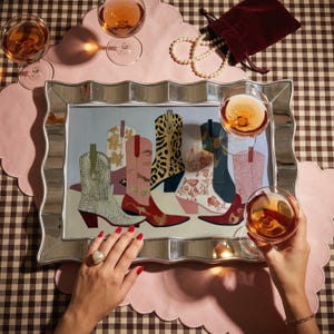 May include: A decorative silver framed artwork featuring a collection of colorful cowboy boots. The boots are painted in various patterns and colors, including leopard print and floral designs. The image is set on a pink scalloped placemat with glasses of rose wine.