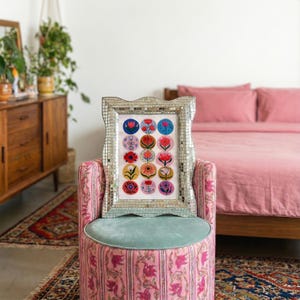 May include: A framed floral art print with a mirrored mosaic frame rests on a pink patterned ottoman with a teal cushion. The print features colorful flower illustrations in circular designs. The background includes a pink bed and a wooden dresser.