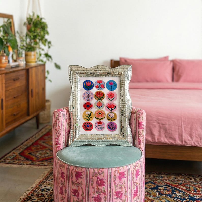 May include: A framed floral art print with a mirrored mosaic frame rests on a pink patterned ottoman with a teal cushion. The print features colorful flower illustrations in circular designs. The background includes a pink bed and a wooden dresser.
