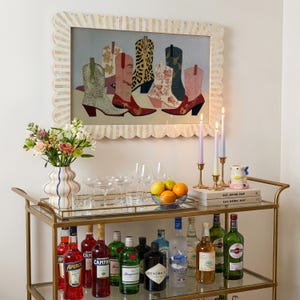 May include: A gold bar cart with various bottles of liquor, glassware, and a vase of flowers. A framed artwork of cowboy boots hangs above the cart. Candles and a small ceramic cup are also on the cart.