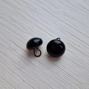 May include: Two glossy black buttons. One is a rounded dome shape with a loop on the back, and the other is a flatter, mushroom-shaped button with a loop. Both buttons are on a light-colored, textured surface.