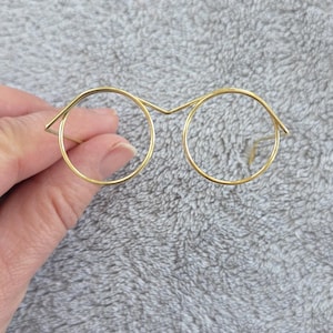 Puede incluir: Delicadas gafas de montura metálica doradas con lentes redondas. Las gafas tienen un diseño minimalista, con monturas finas y un puente que conecta las dos lentes. La estética general es simple y elegante.