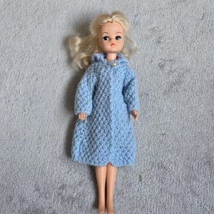 May include: A vintage doll with blonde hair wearing a light blue, hooded, knitted dress. The doll has a classic face with painted features and is posed on a grey, textured surface. A collectible toy.