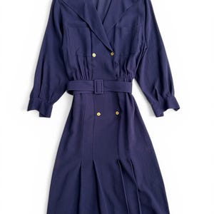 Op de afbeelding: Marineblauwe jurk met een brede kraag, lange mouwen en een ceintuur in de taille. De jurk heeft goudkleurige knopen en een geplooide rok. Het ontwerp omvat twee voorzakken en schouderaccenten. De jurk is een vintage stijl.
