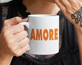 May include: A white ceramic mug with a black handle and the word "AMORE" printed in orange on the front. The mug is being held by a person with a tattoo on their hand.
